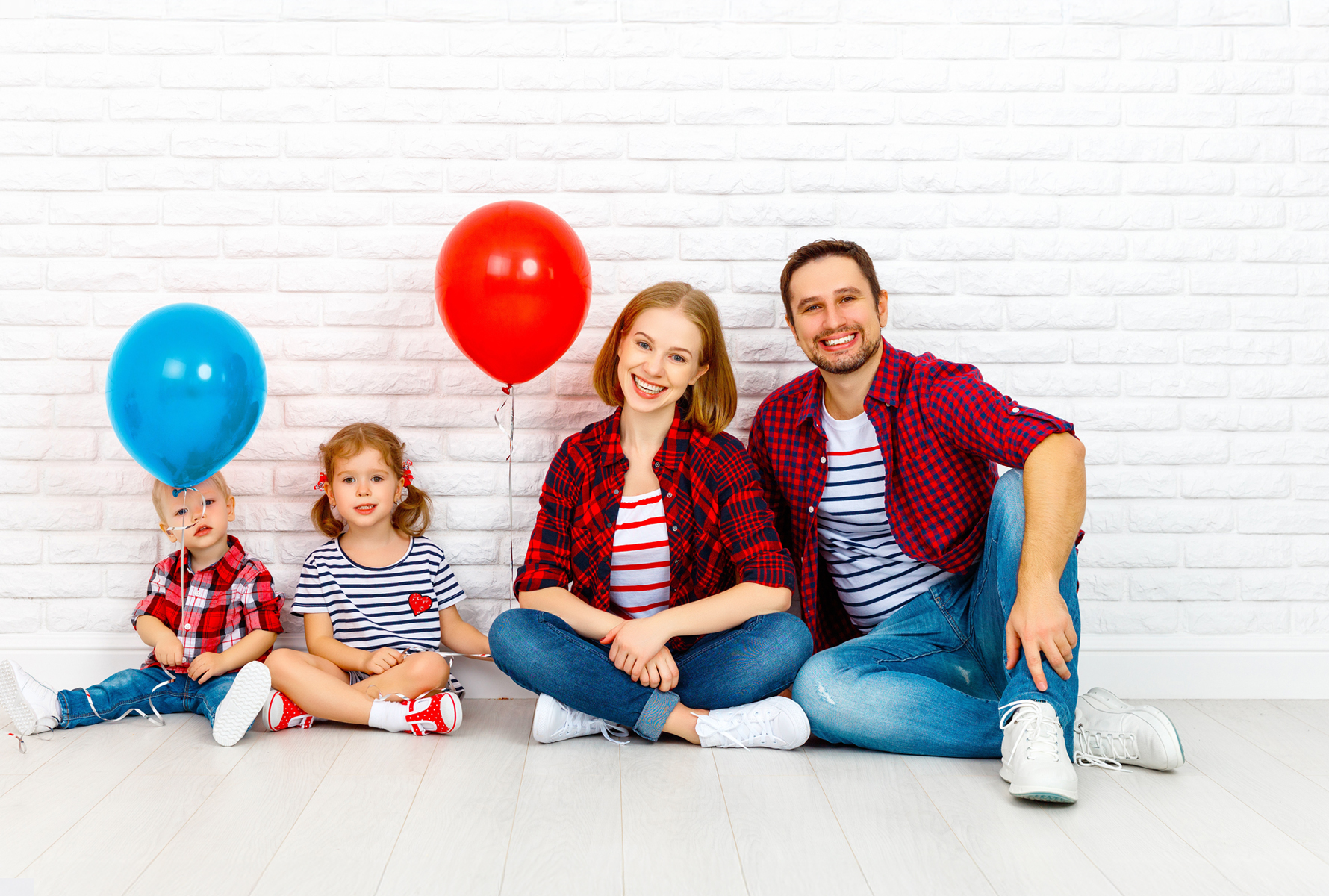 Familie mit Ballons vor Backsteinmauer Die gleiche Familie sitzt entspannt vor einer weissen Backsteinmauer, die Kinder halten einen roten und blauen Ballon – ein Moment voller Leichtigkeit.