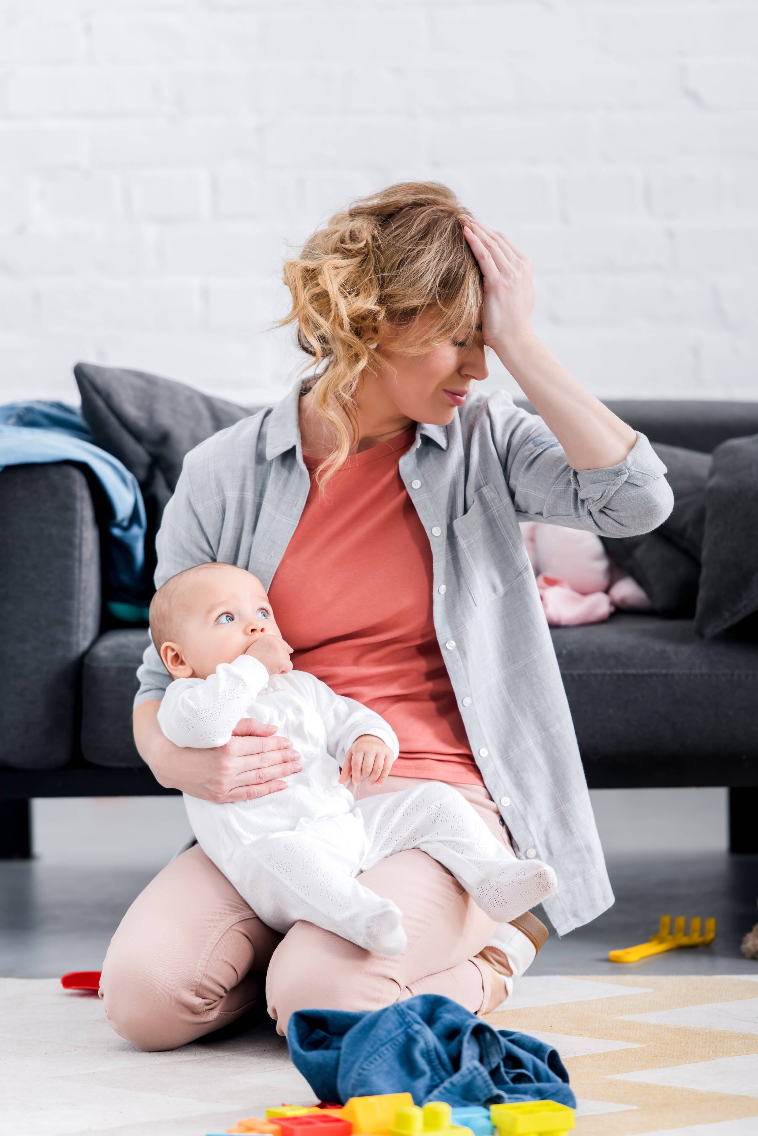 Überforderung im Alltag – Mutter mit Kind Das Bild zeigt eine Mutter mit ihrem Baby auf dem Boden sitzend, umgeben von Spielsachen und Alltagsgegenständen. Es vermittelt eine Situation der Überforderung und den Bedarf an Unterstützung im Familienalltag.
