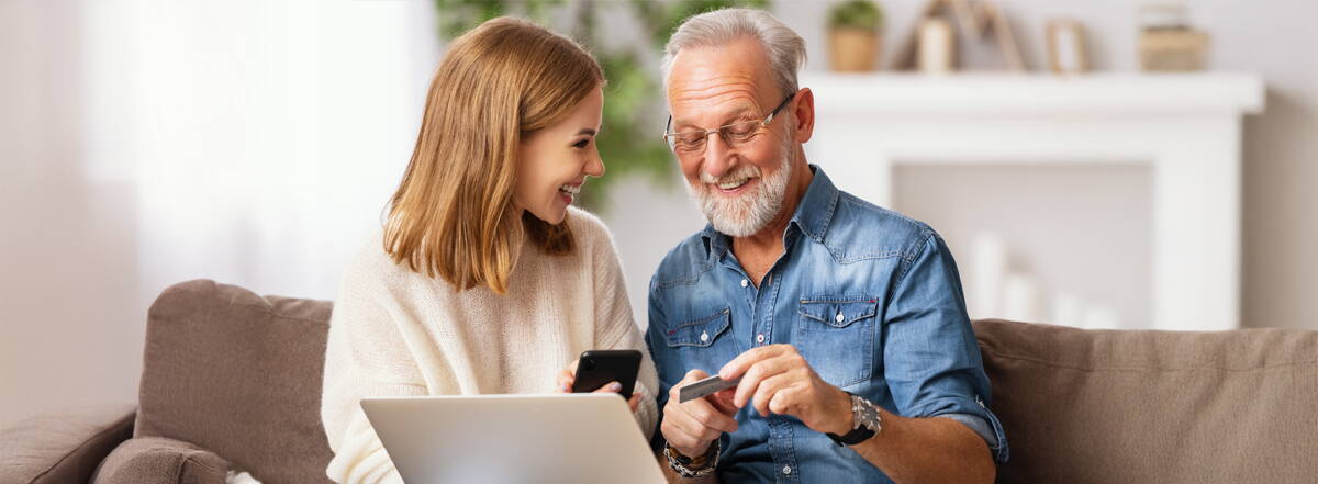 Kundenfreundlich versichert Eine junge Frau und ein älterer Herr sitzen gemeinsam auf dem Sofa. Laptop, Handy und Bankkarte zeigen: Hier geht Beratung auf Augenhöhe.