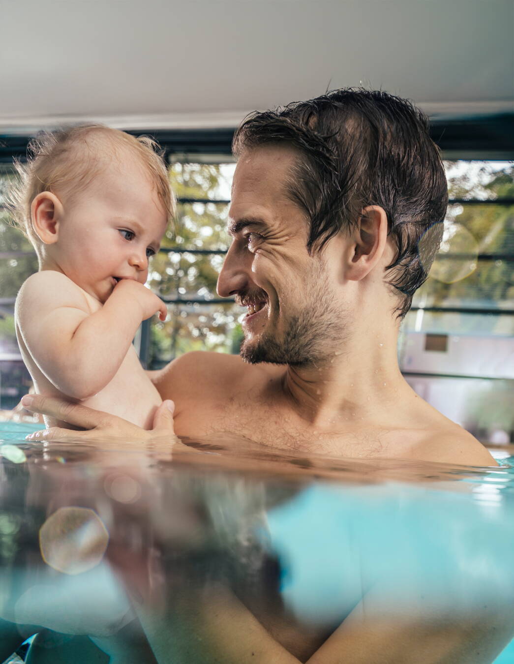 Père et enfant dans un cours de natation pour bébés Un homme tient son jeune enfant dans les bras, tous deux rient dans l’eau chaude d’une piscine couverte – proximité et confiance grâce à la natation pour bébés.