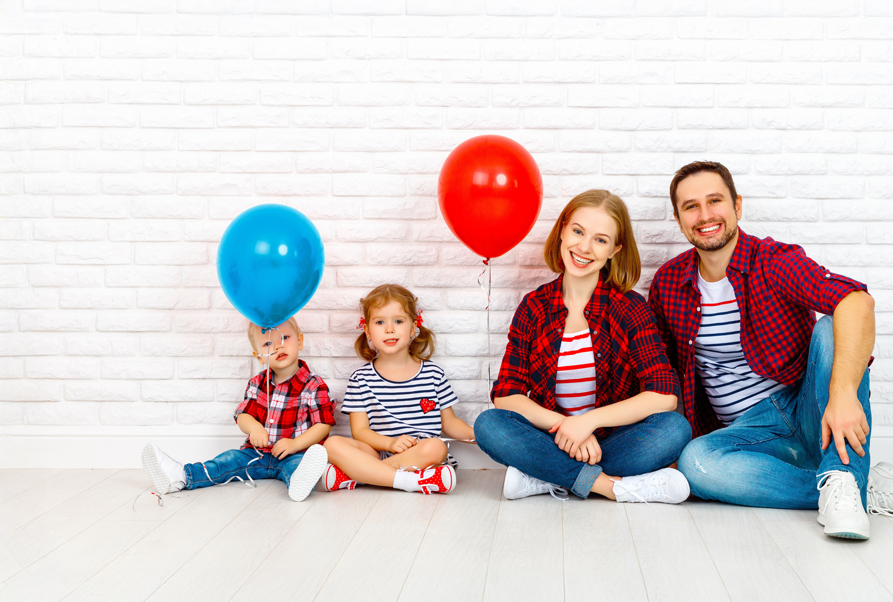 Une famille composée d’un père, d’une mère et de deux enfants sourit à la caméra – symbole de cohésion et de joie de vivre. La même famille est assise détendue devant un mur en briques blanches, les enfants tiennent un ballon rouge et un ballon bleu – un moment plein de légèreté.