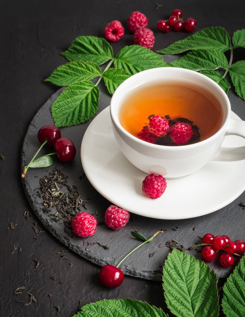 Tisane de feuilles de framboisier dans une tasse blanche posée sur une ardoise Une tasse de tisane de feuilles de framboisier avec deux framboises fraîches, servie sur une ardoise. À côté se trouvent des feuilles de framboisier fraîches et séchées ainsi que des framboises.
