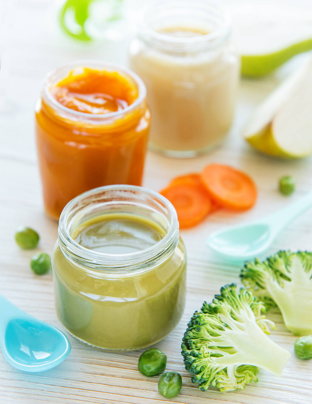 Un inizio variopinto con le pappe Tre vasetti di pappa fatta in casa sono disposti su un tavolo di legno chiaro, accanto a cucchiaini per bebè, broccoli, piselli, carote e una mela – tutto è pronto per le prime cucchiaiate di divertimento
